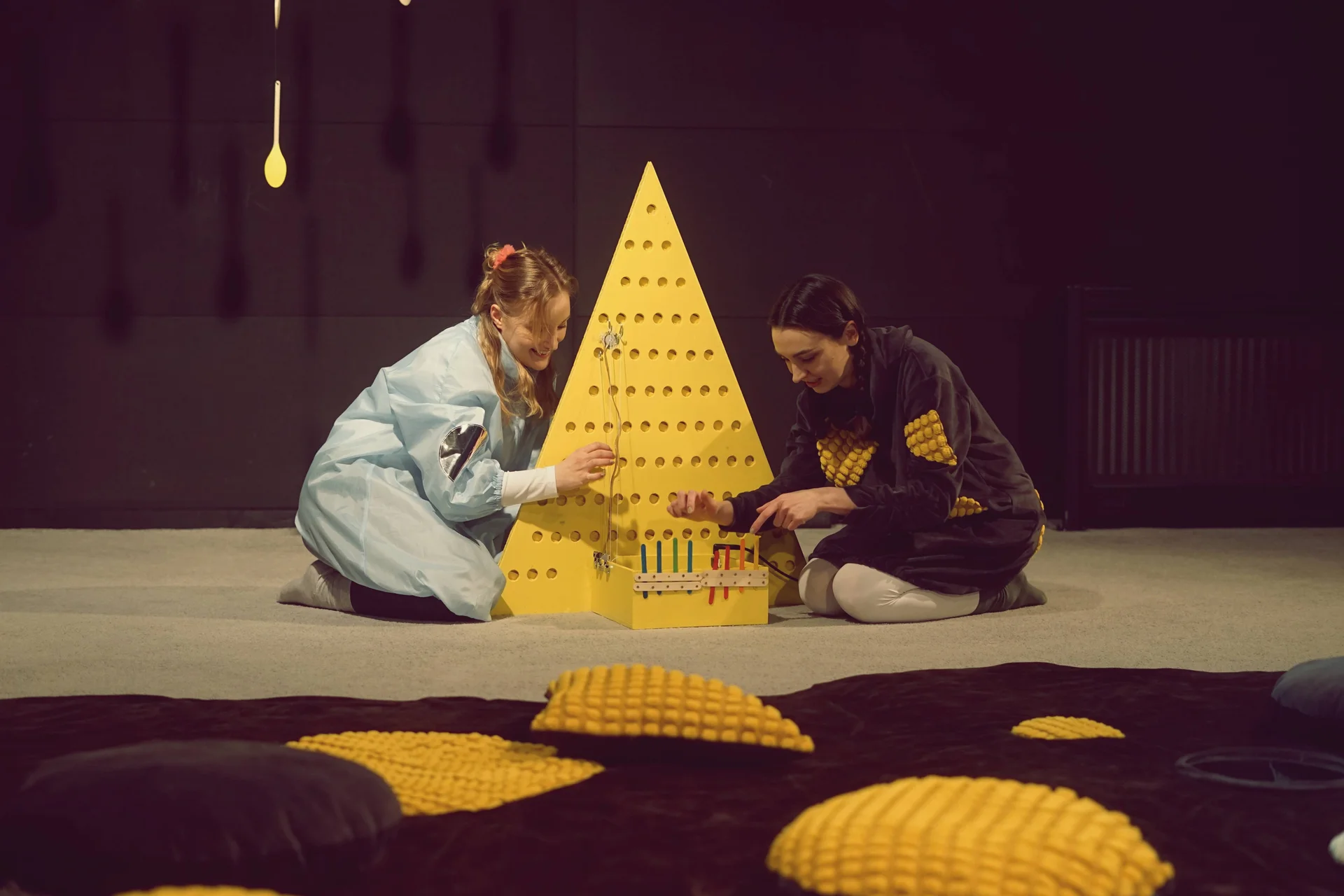 dwie aktorki animujące przedmiotami przed żółtym elementem scenografii w kształcie trójkąta